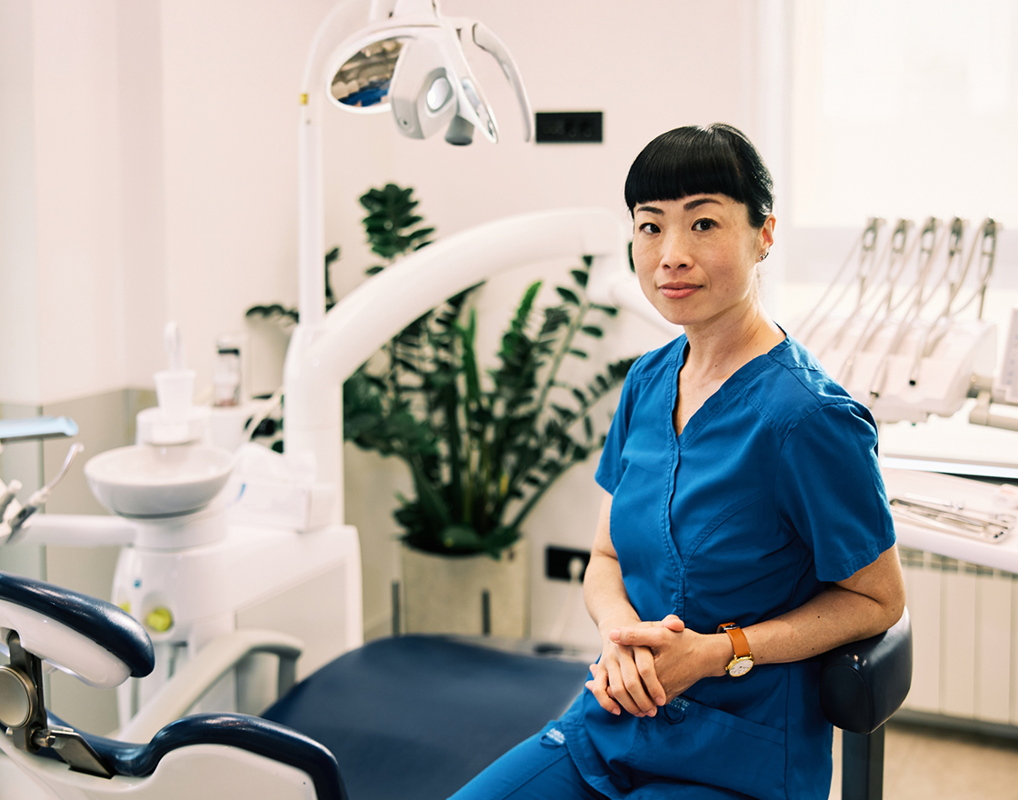 Woman in blue scrubs seated on a stool beside a dental chair in a bright clinic, hands folded, with an overhead light, dental instruments, and a potted plant visible in the background.
