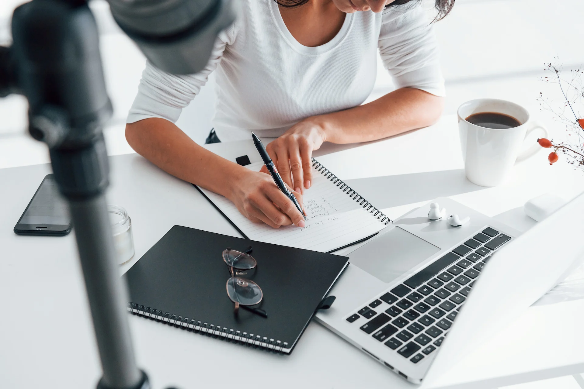 Hands of a person writing in a spiral notebook at a bright white desk, with a laptop, a black notebook topped by glasses, a smartphone, wireless earbuds, and a coffee mug nearby and a camera tripod blurred in the foreground.