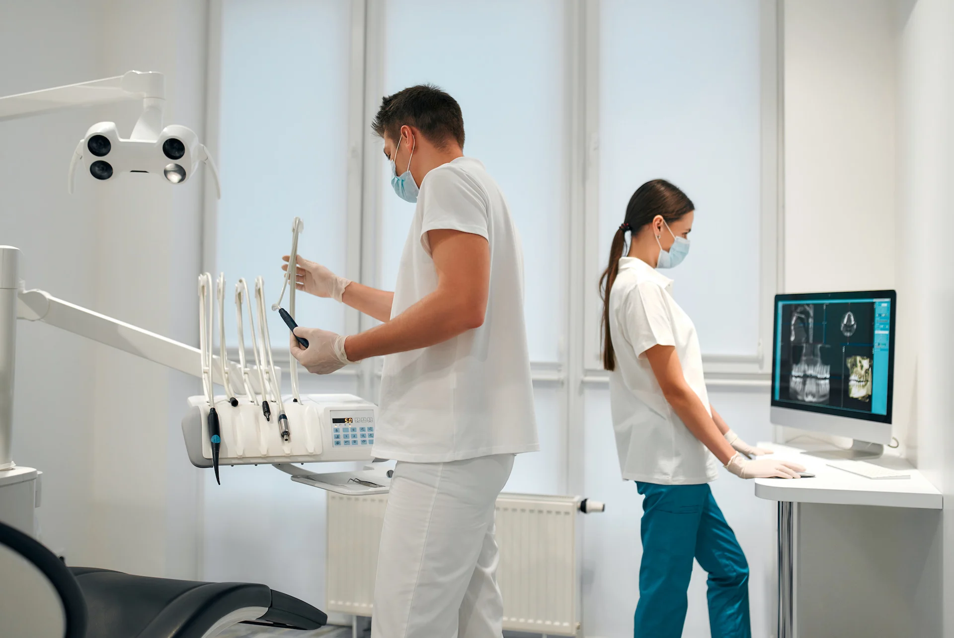 Two masked dental professionals in a bright operatory—one adjusting dental handpieces by the chair and overhead light, the other reviewing dental X‑ray images on a computer monitor.