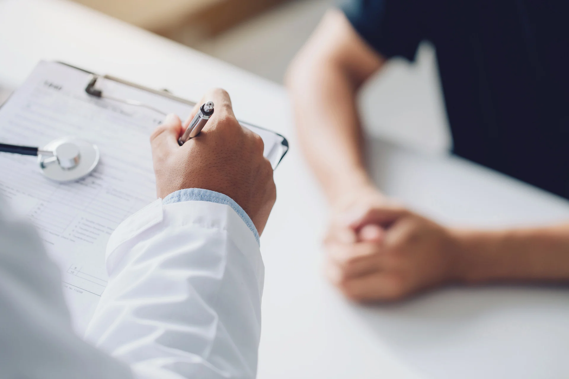 Doctor's hand holding a pen and filling out a clipboard beside a stethoscope, with a patient's folded hands blurred in the background.