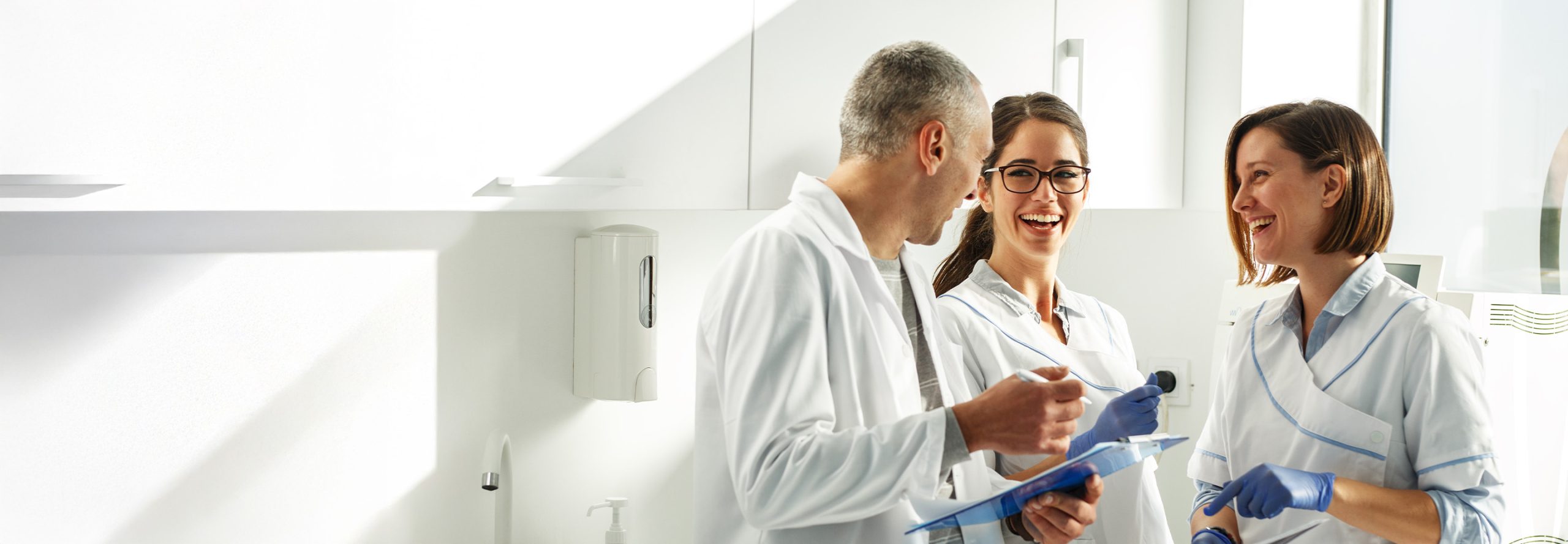 Three medical staff—two women and one man—in white uniforms and blue gloves laugh and discuss notes on a clipboard in a bright, white clinical room.