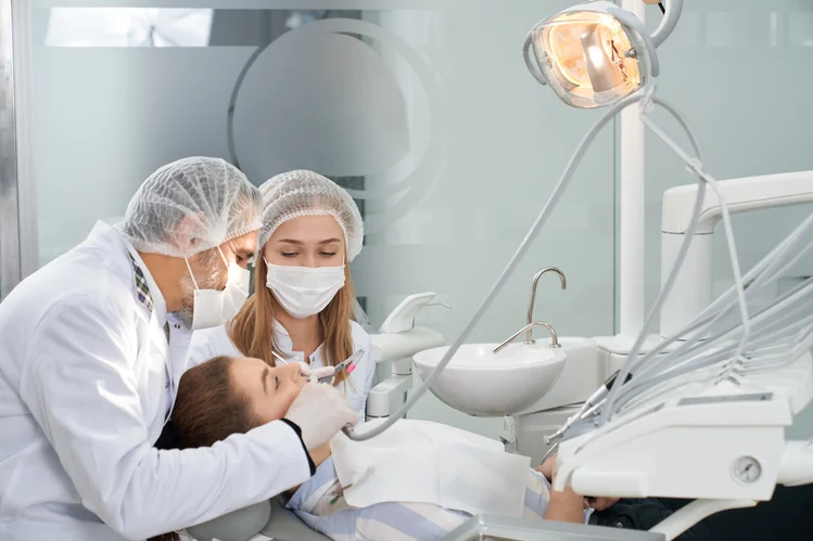 Dentist and dental assistant in masks and hairnets work on a child reclined in a dental chair, with dental instruments, an illuminated overhead lamp, and a sink visible in the treatment room.