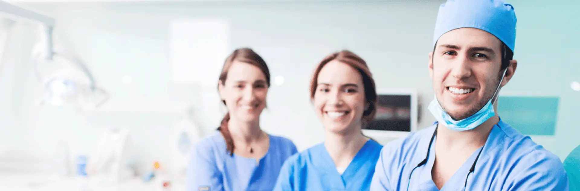 Three smiling healthcare professionals in blue scrubs in a bright clinic, with a male surgeon in a blue cap and mask pulled down in the foreground and two female colleagues blurred behind him.