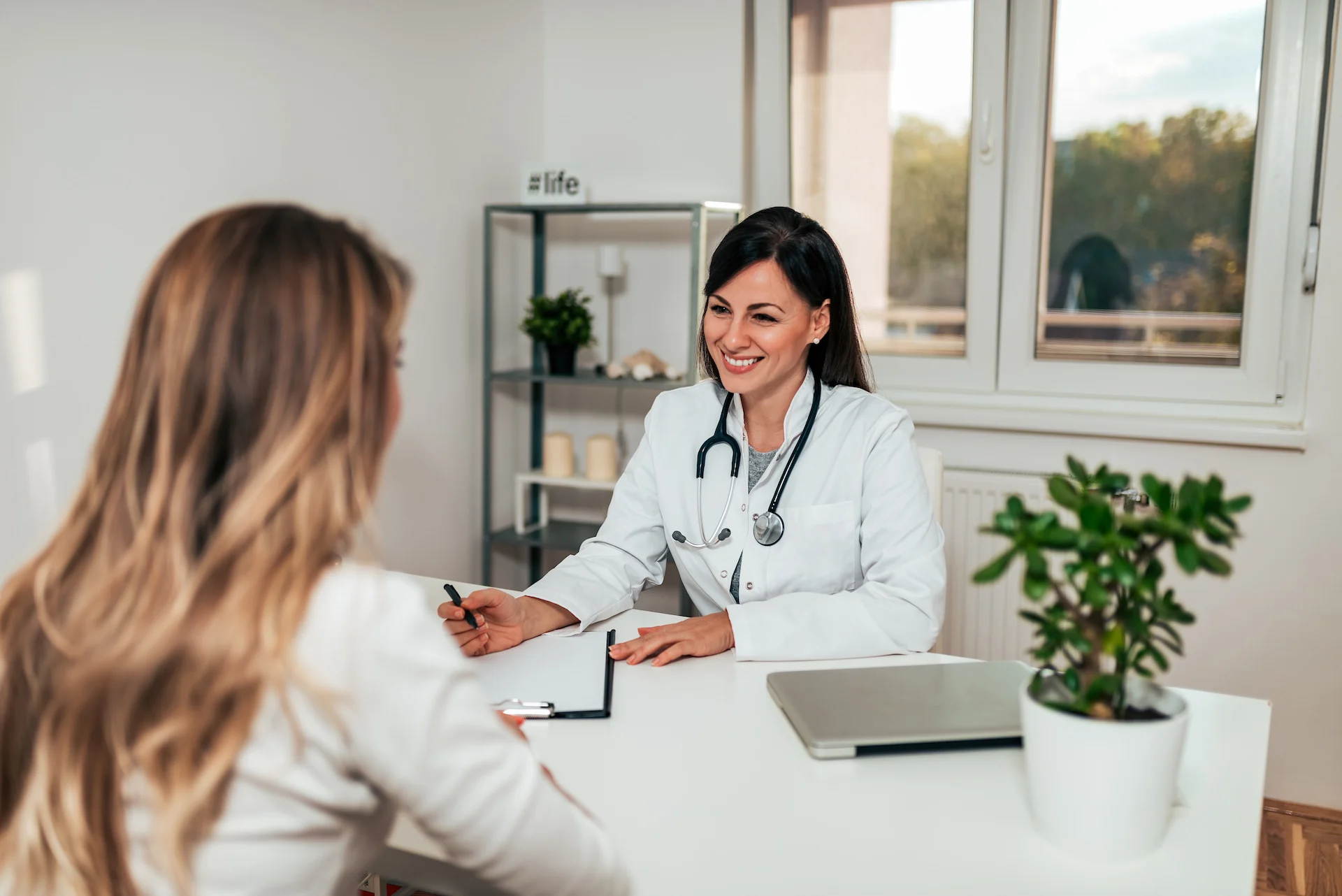 A smiling female doctor in a white coat with a stethoscope consults a patient across a desk with a clipboard, laptop and potted plant in a bright medical office with shelving and a window.