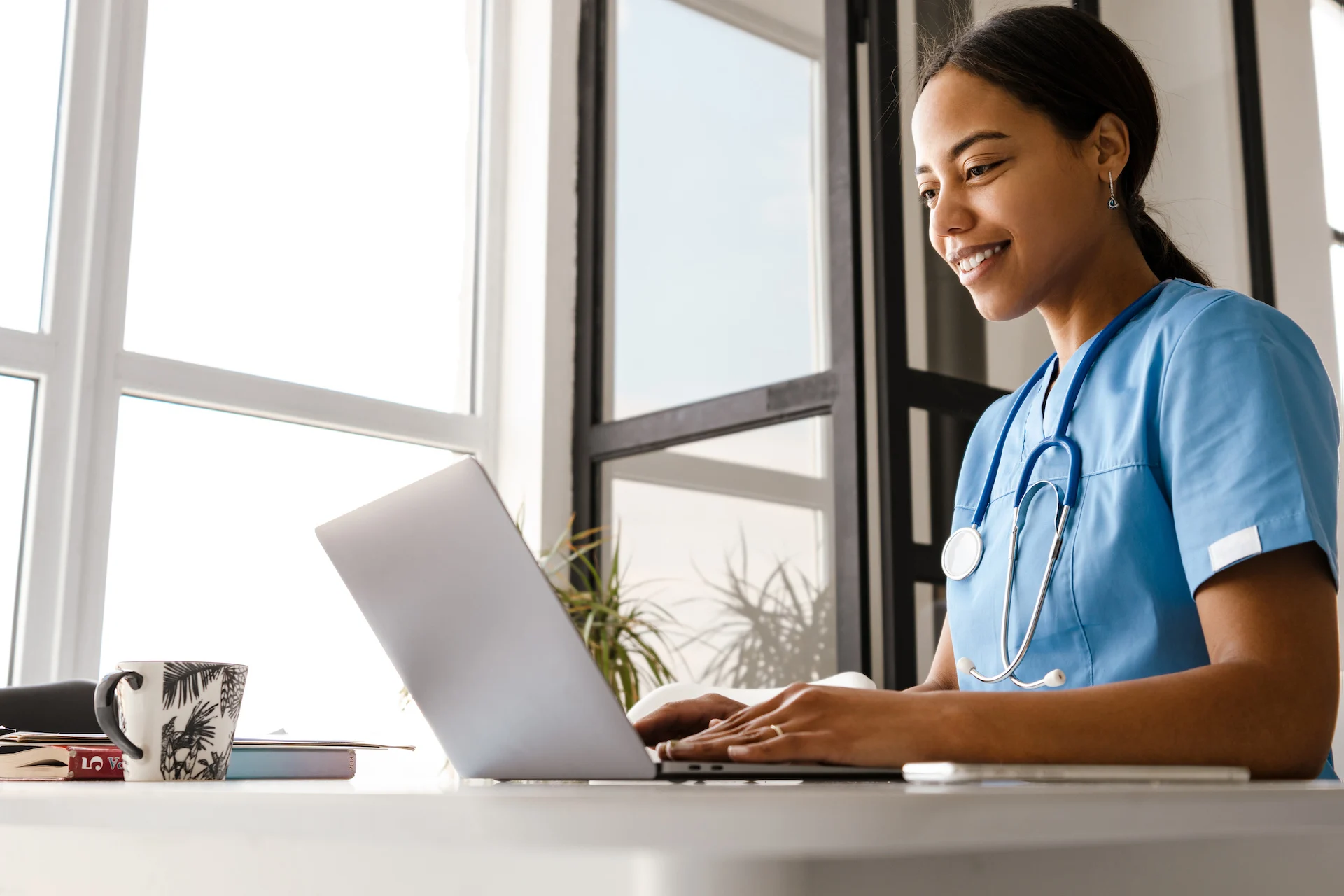 Smiling nurse in blue scrubs with a stethoscope around her neck types on a laptop at a bright desk by a window, with a patterned coffee mug and notebooks nearby.