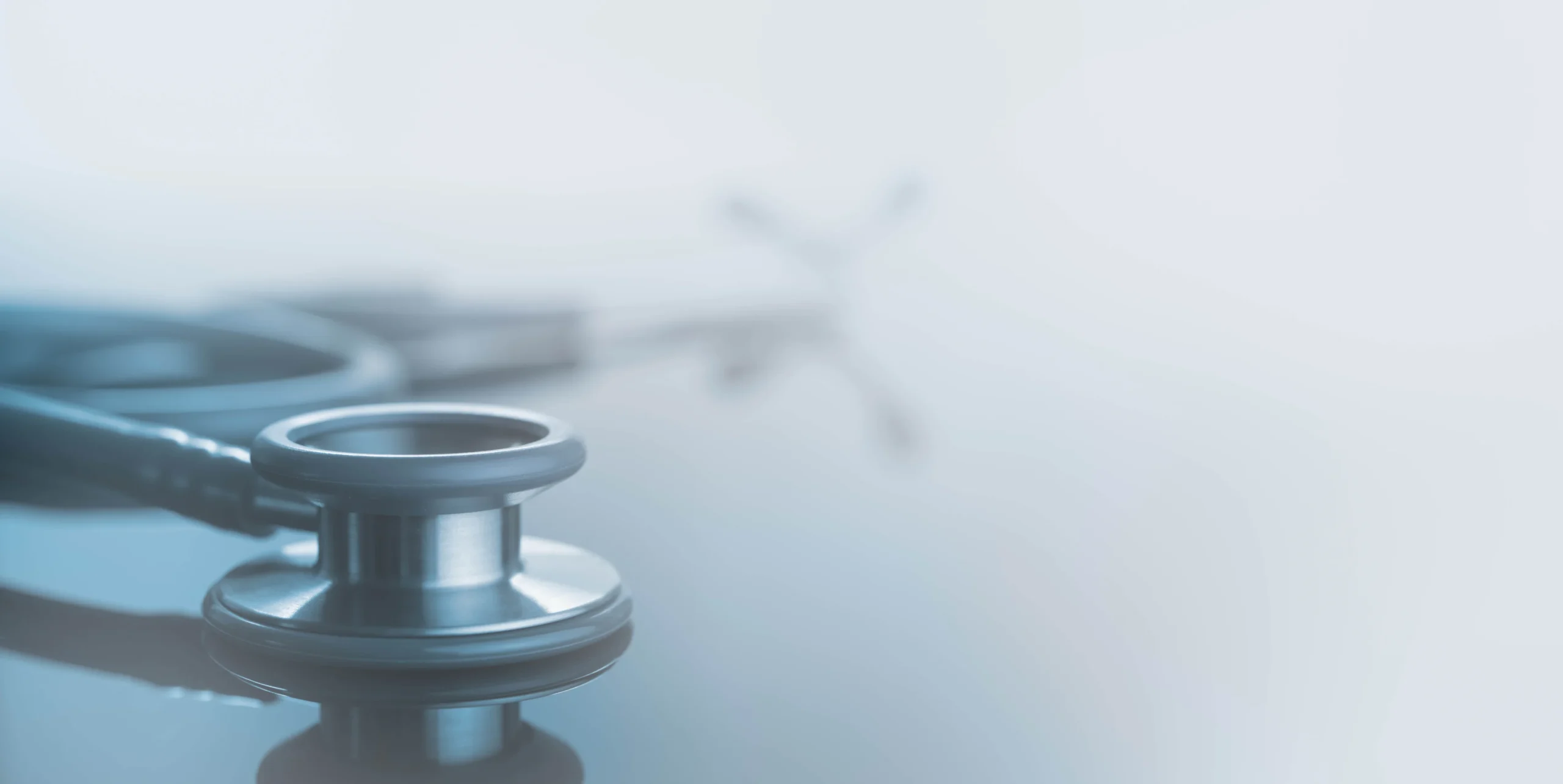Close-up of a stethoscope chestpiece resting on a reflective surface with coiled tubing and blurred earpieces in soft blue lighting.