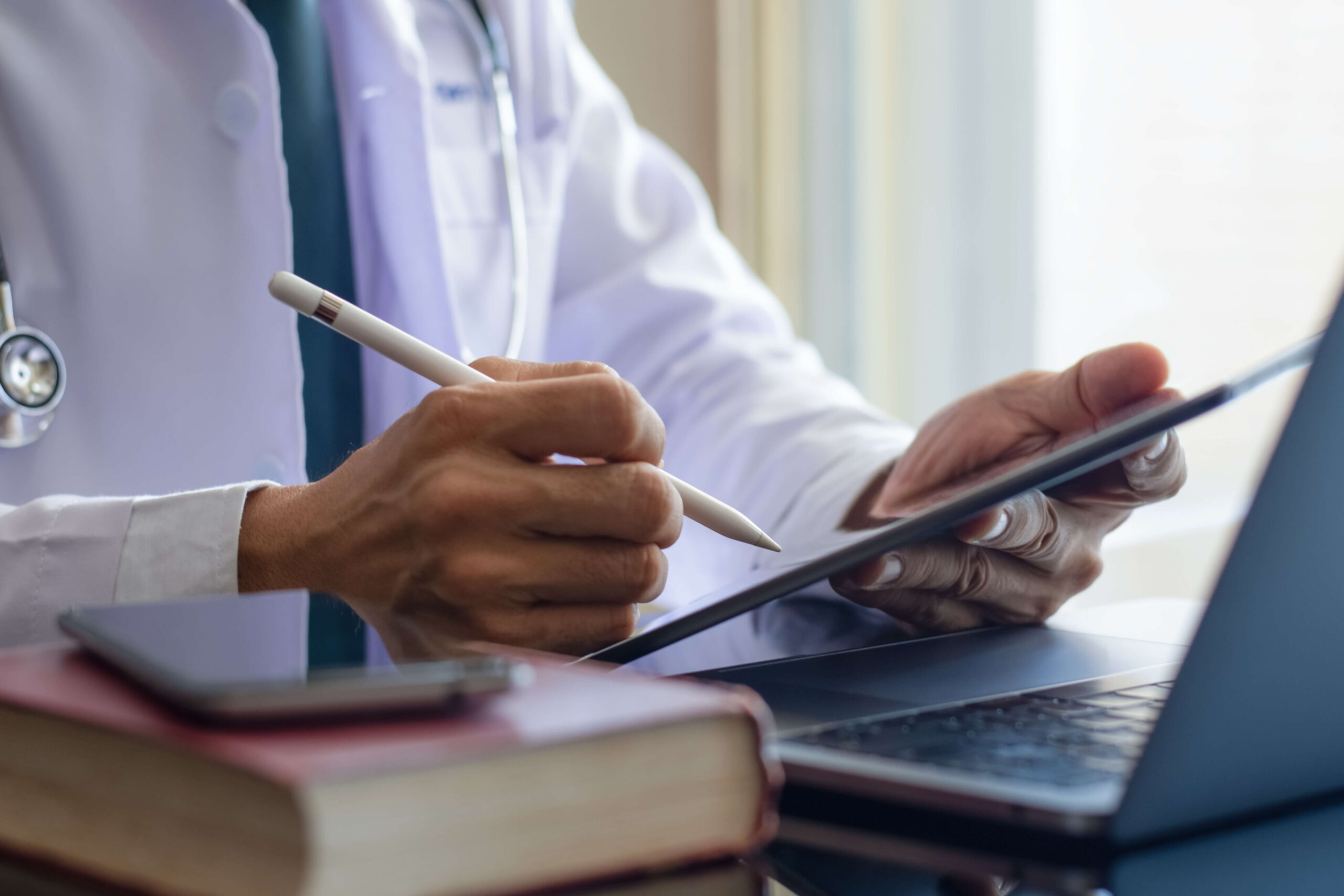 Physician's hands using a stylus on a tablet beside a laptop, with a white coat, stethoscope and a book on the desk.