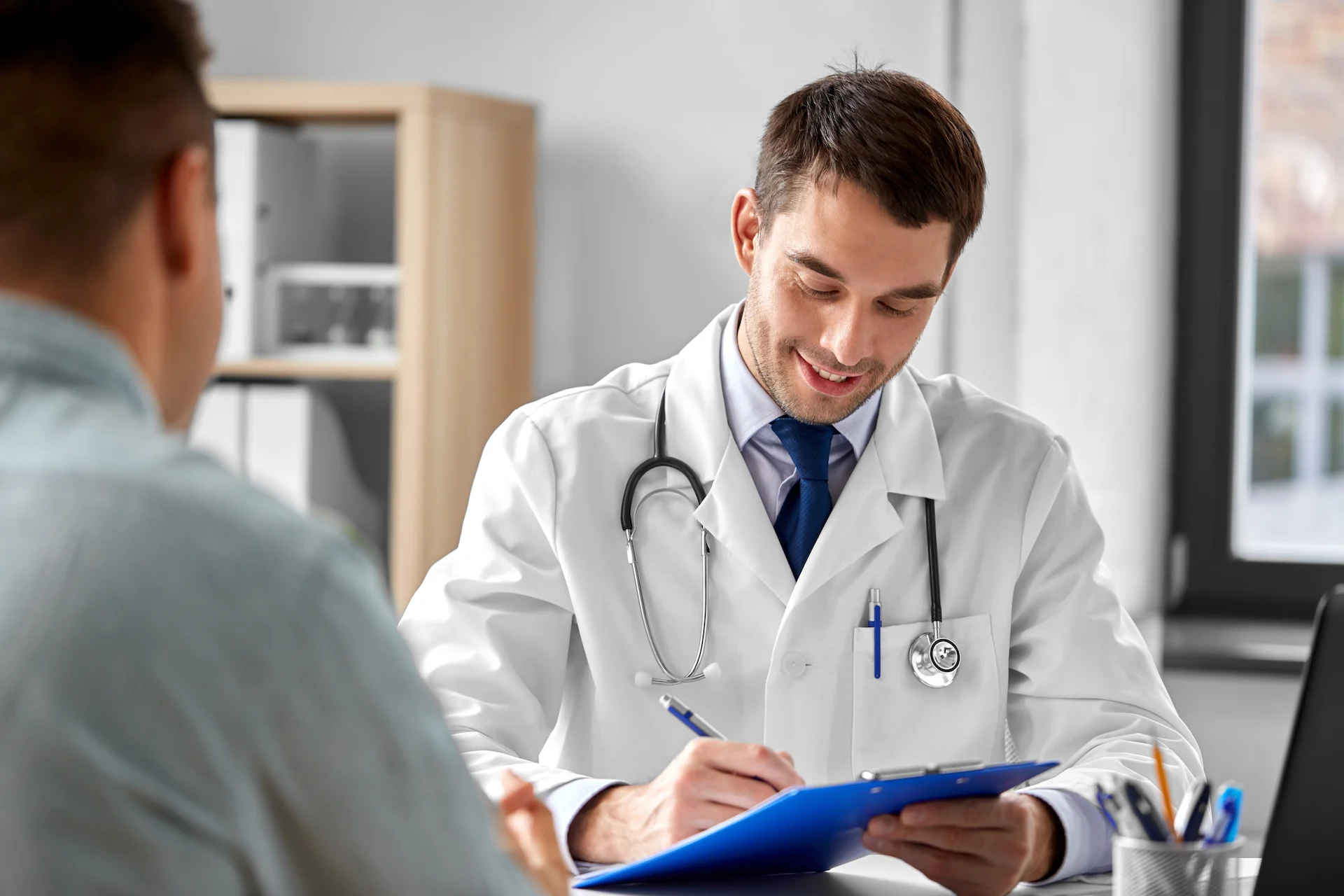 Smiling male doctor in a white coat with a stethoscope around his neck writing on a blue clipboard while consulting a patient in a bright office with shelves and a window.