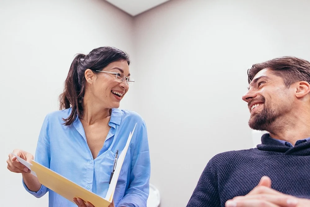Two colleagues smiling at each other in a bright office: a woman in glasses and a light blue blouse holds a yellow folder and pen while a man in a dark sweater sits facing her.