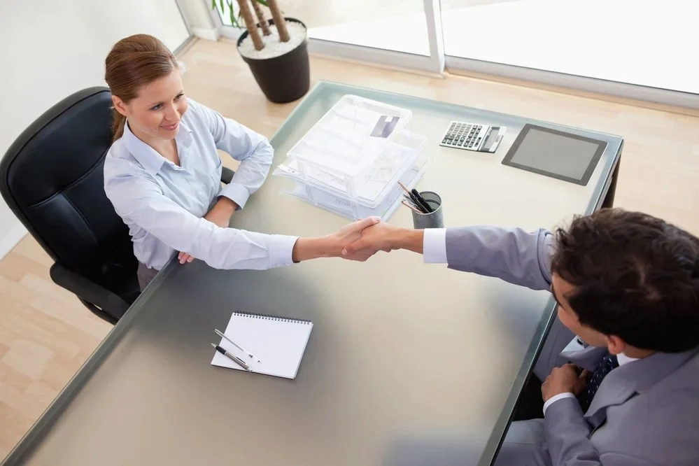 Two businesspeople shaking hands across a glass office desk, the woman seated on the left smiling and the man on the right, with a notepad and pens, document trays, a tablet, a calculator on the desk and a potted plant by the window.