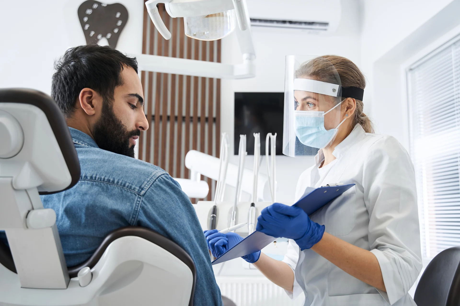 Dentist in a face shield, surgical mask and blue gloves speaks to a male patient in a denim jacket seated in a dental chair, holding a clipboard and pen with dental instruments and lights in the bright clinic behind them.