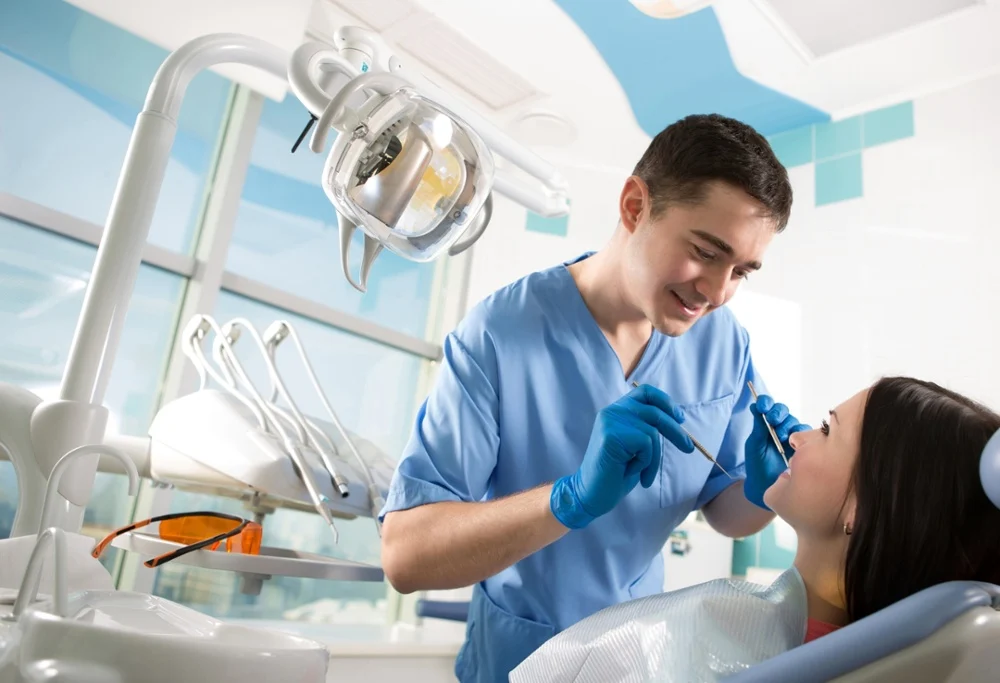 Male dentist in blue scrubs and gloves leans over a female patient in a dental chair holding a dental mirror and probe in a bright modern clinic with an overhead light and instruments nearby.