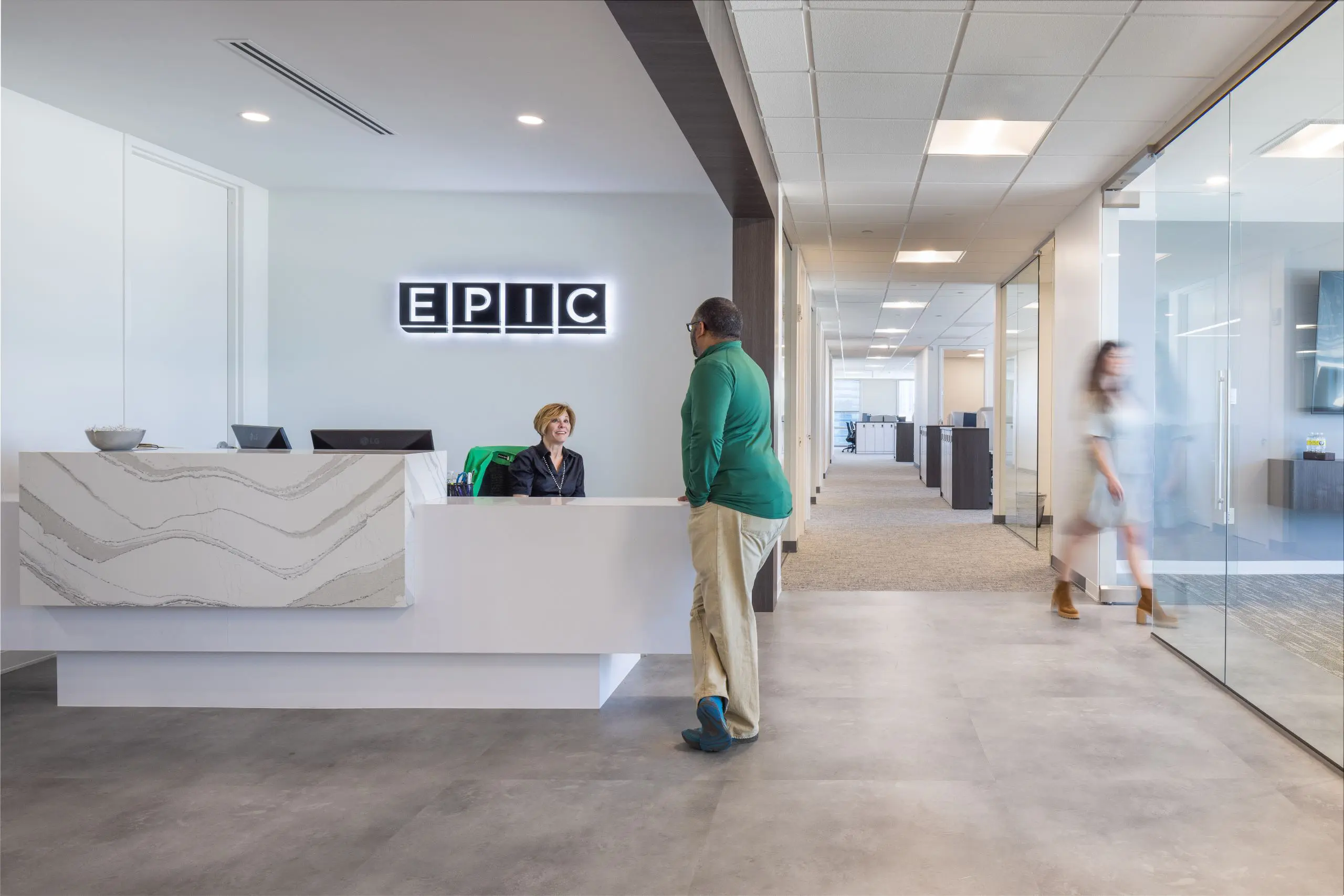 Modern office reception with a white marble-front desk under an illuminated 'EPIC' wall sign, a smiling receptionist seated behind the desk speaking with a man leaning on the counter and a glass-walled corridor to the right where a woman walks by in motion blur.