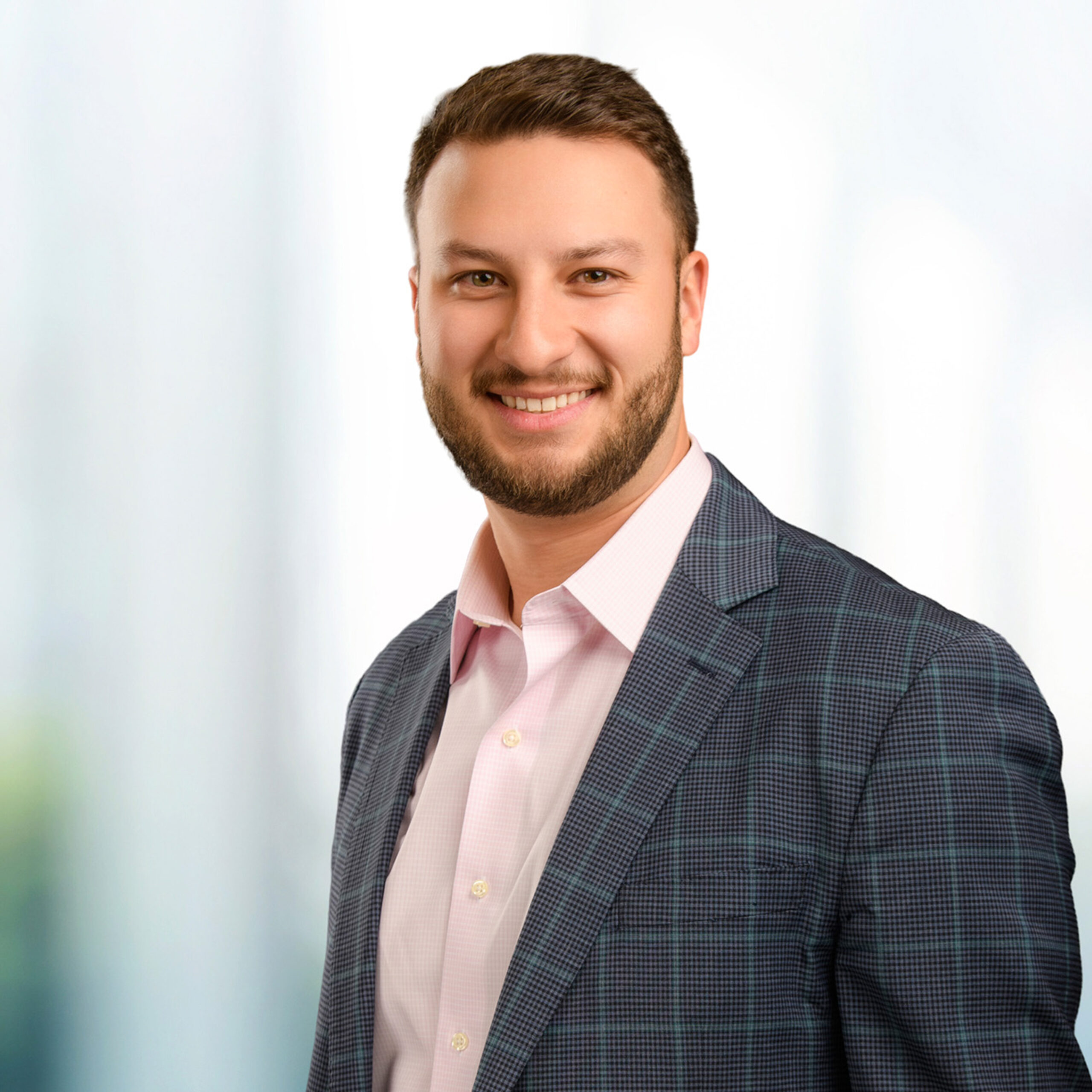 Smiling bearded man wearing a light pink dress shirt and blue checked blazer against a soft blurred background.