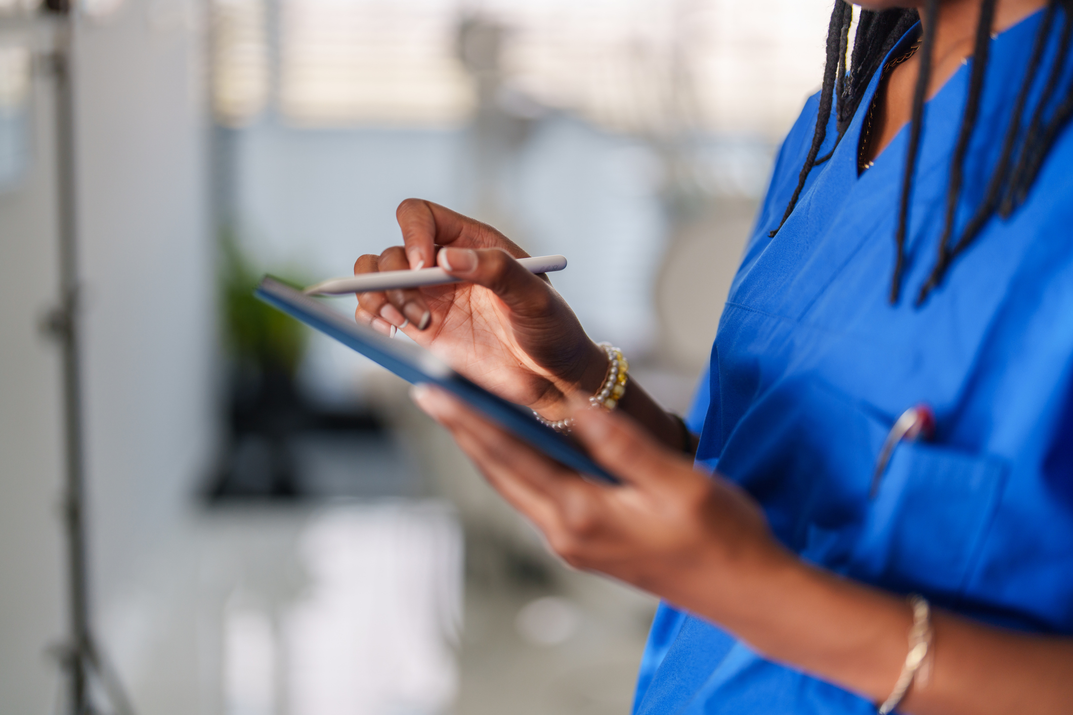 Close-up of a healthcare worker in blue scrubs holding a tablet and tapping it with a stylus, with braided hair, bracelets on the wrist, and a stethoscope partly visible in a pocket.