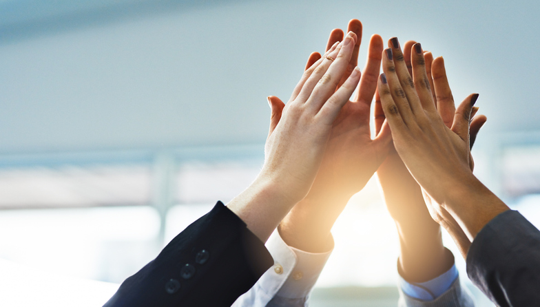 Several people in business attire joining hands in a group high-five above their heads with sunlight streaming between them