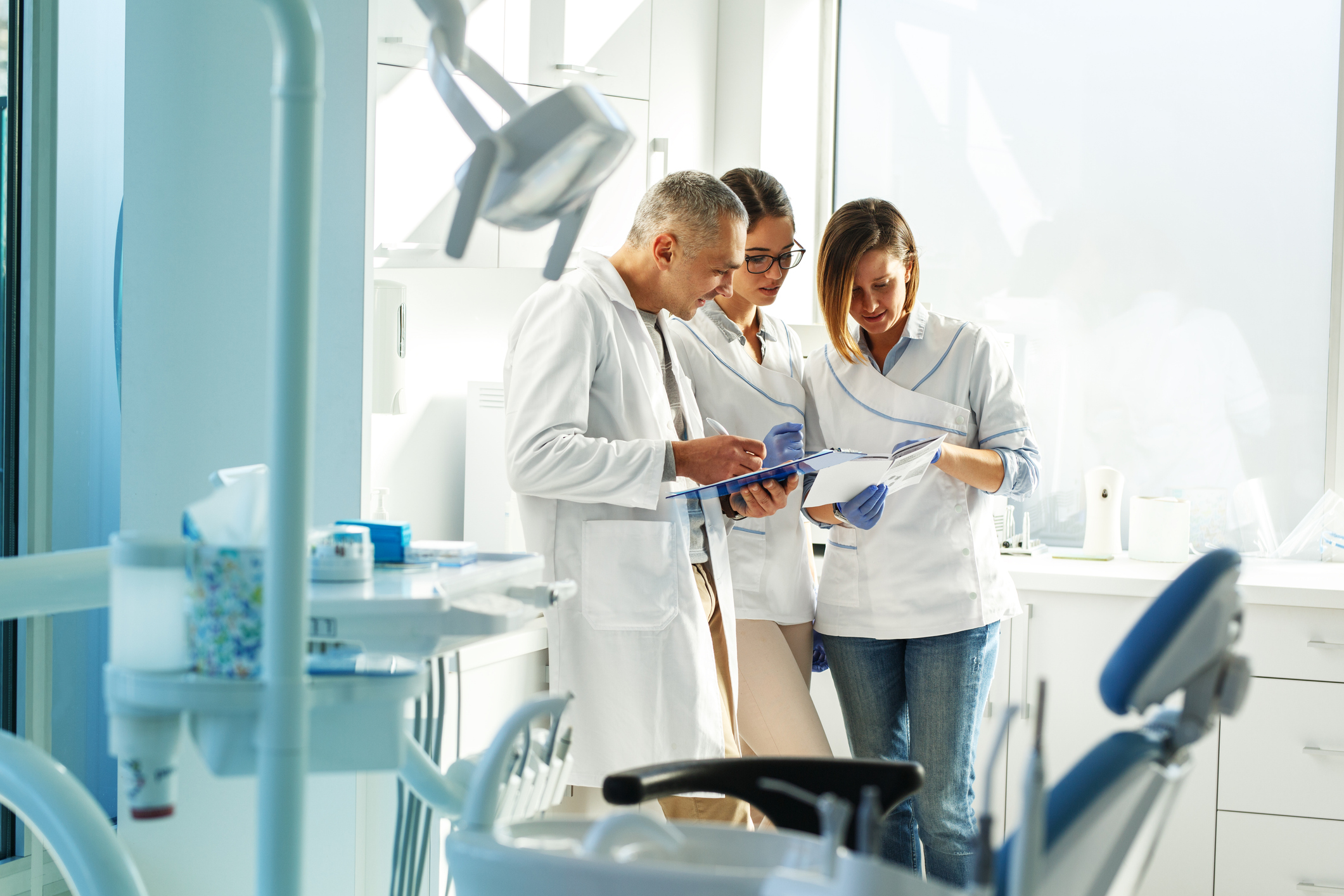 Three dental staff wearing white coats and gloves stand in a bright dental operatory, reviewing patient charts on a clipboard beside a dental chair and overhead lamp.