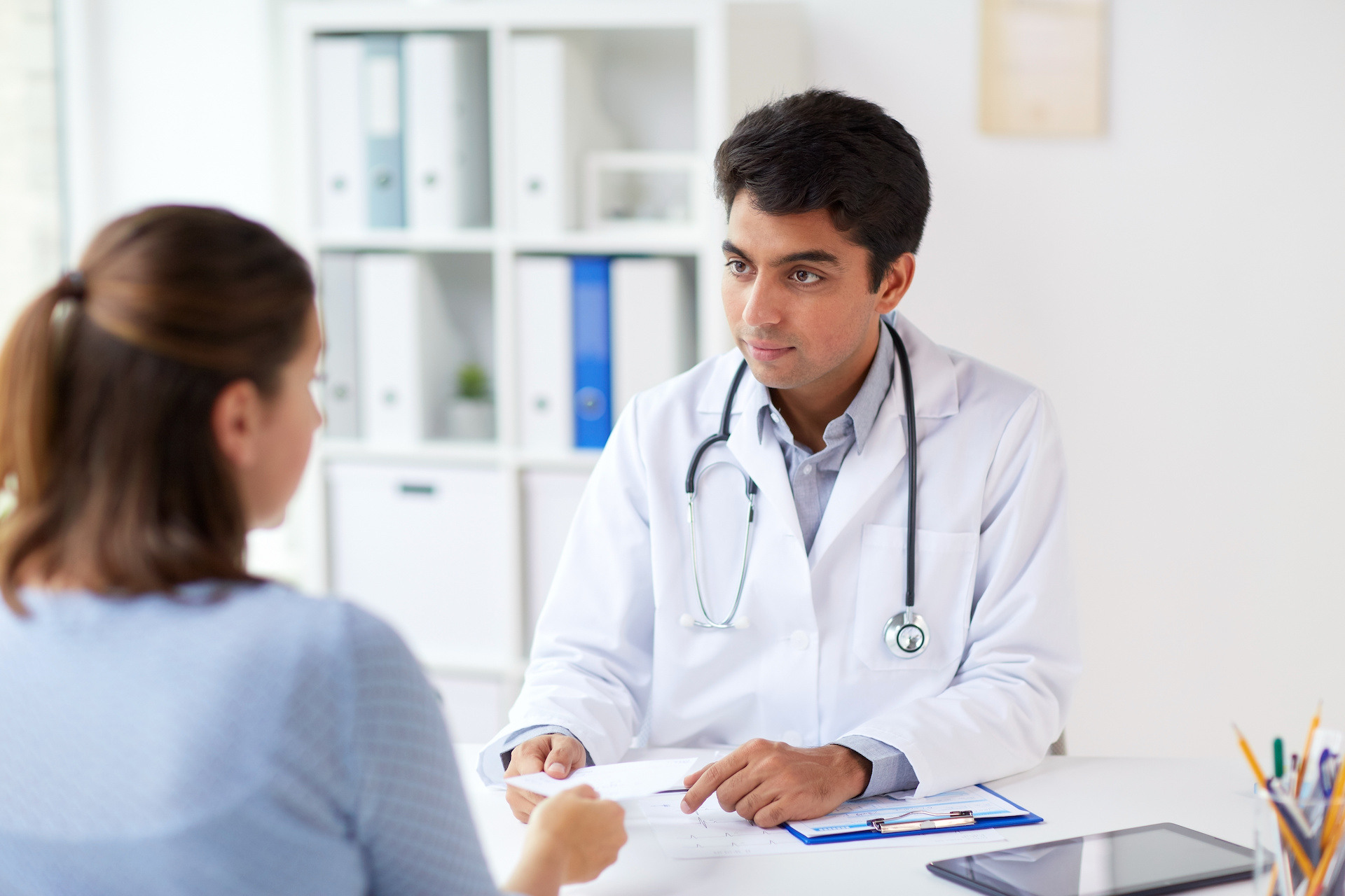 Doctor in a white coat with a stethoscope sits at a desk in a bright medical office, attentively speaking to a seated patient and handing over a document while a clipboard and tablet rest on the desk.