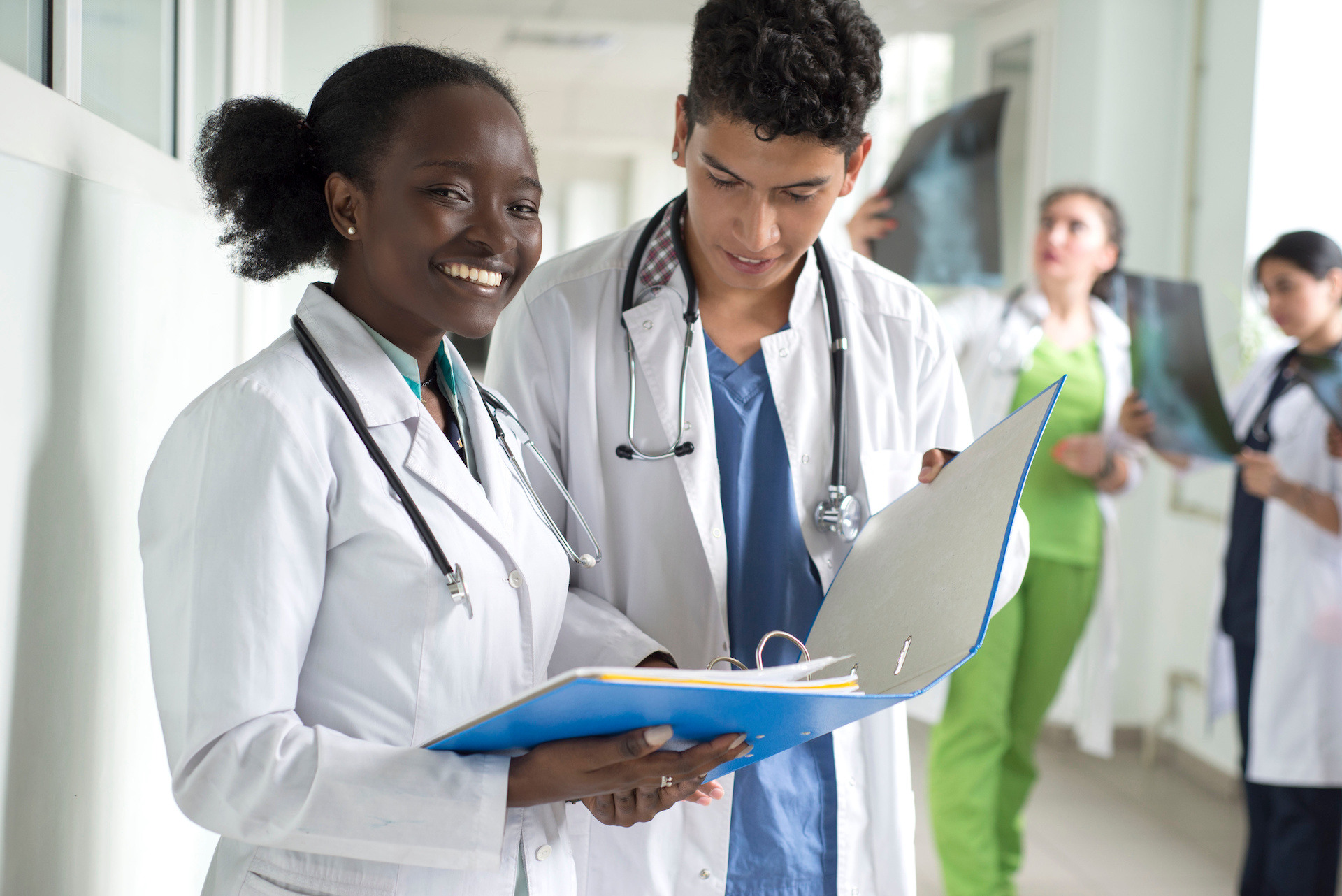 Two doctors in white coats and stethoscopes reviewing a blue patient file in a bright hospital corridor, one smiling at the camera while colleagues examine X-rays in the background.